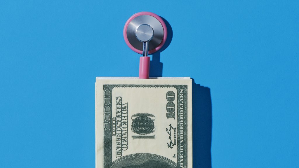 An Expert's Take on Long-Term Care Insurance: Buy or Not? A pink stethoscope and a pile of hundred-dollar bills against a blue background.