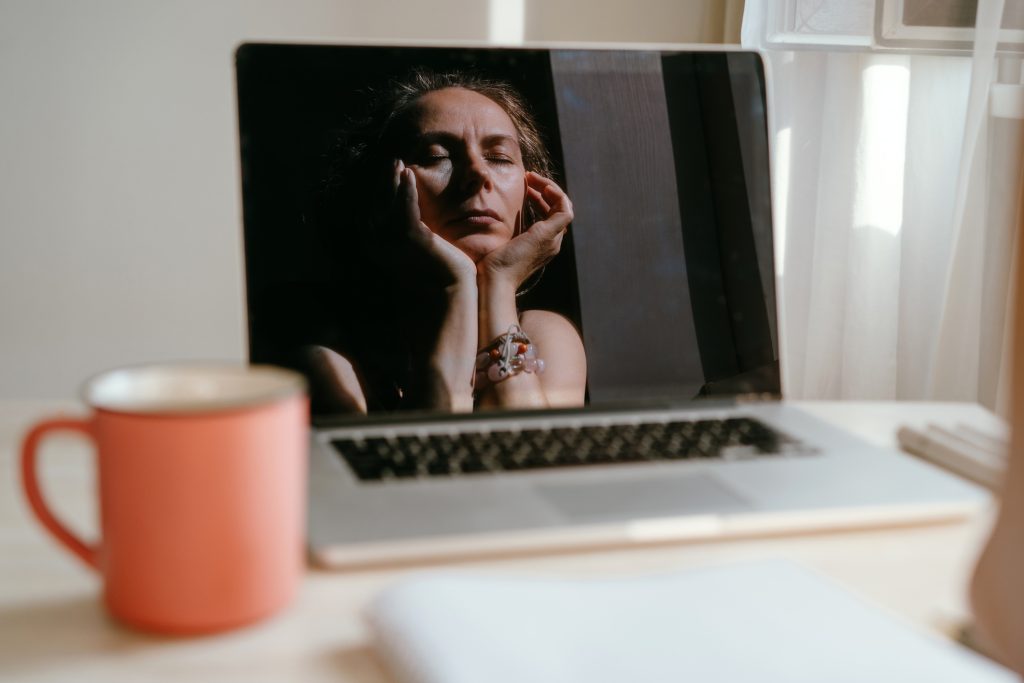 Portrait of a woman sitting at the notebook, red cup of tea on the table. Reflection of a face in the monitor screen, chin on hands. She looks tired.