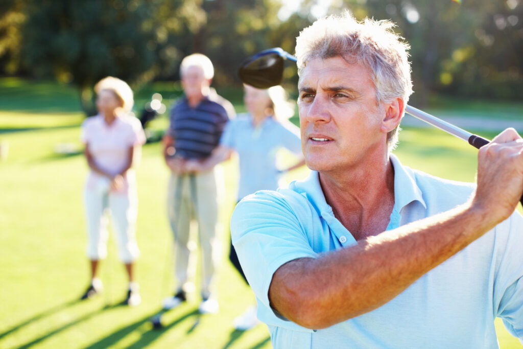 Back to Golf School — for Adults and Retirees Handsome, mature man swinging his golf club with friends in blurred background