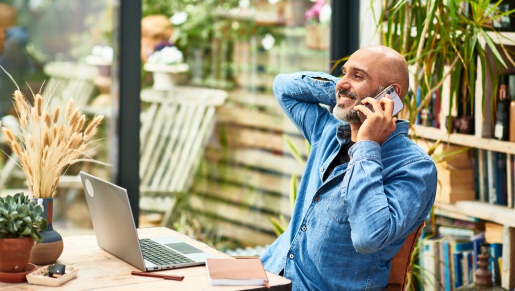 A mature man sits in his home office on a cellphone with a laptop in front of him.