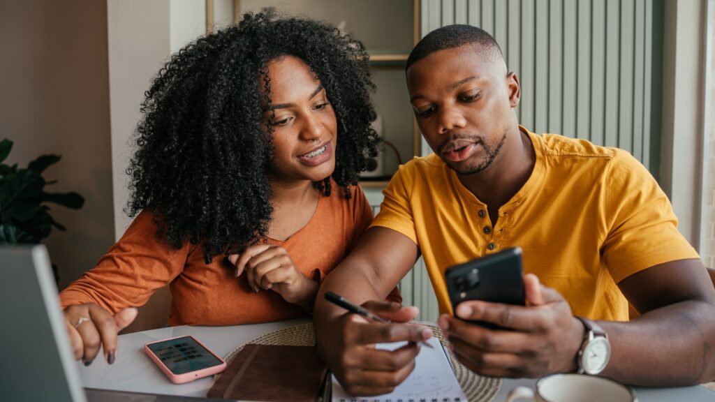 Four Ways You Can Use Debt to Build Wealth A young couple look at their phones while financial planning at their dining room table.