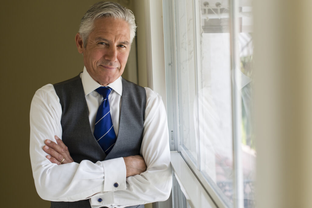 I'm 57 With $4.1 Million and Looking to Retire Abroad in a Few Years. I No Longer See the Point in Contributing to My 401(k). Am I Wrong? A wealthy-looking businessman in his 50s poses next to a window with his arms crossed.