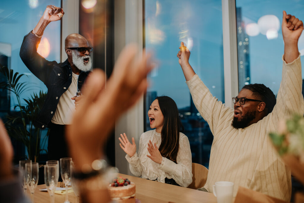 Three people who are gathered around a table with cake and champagne glasses and are cheering.