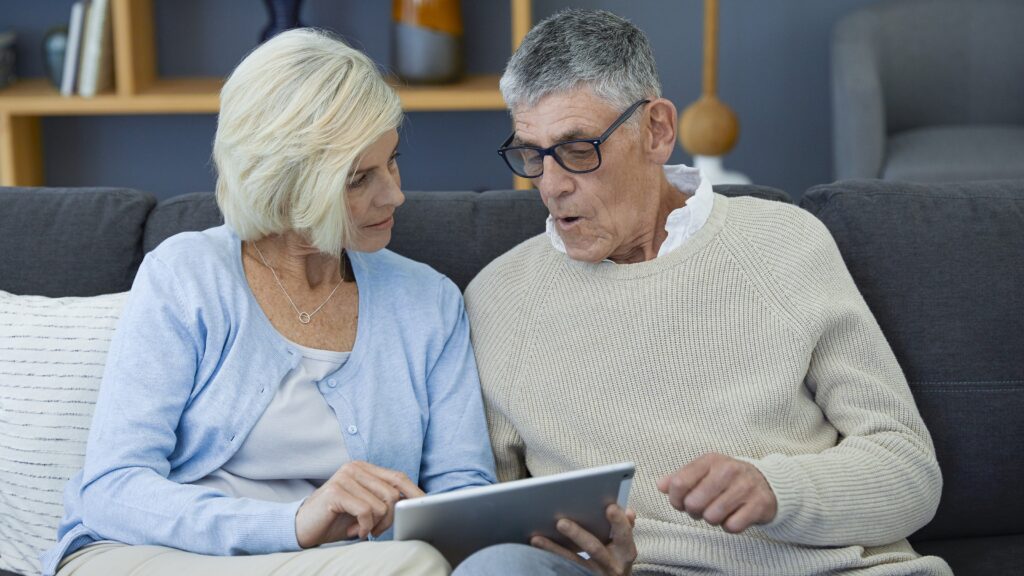 An older couple look at a tablet together while sitting on their sofa.