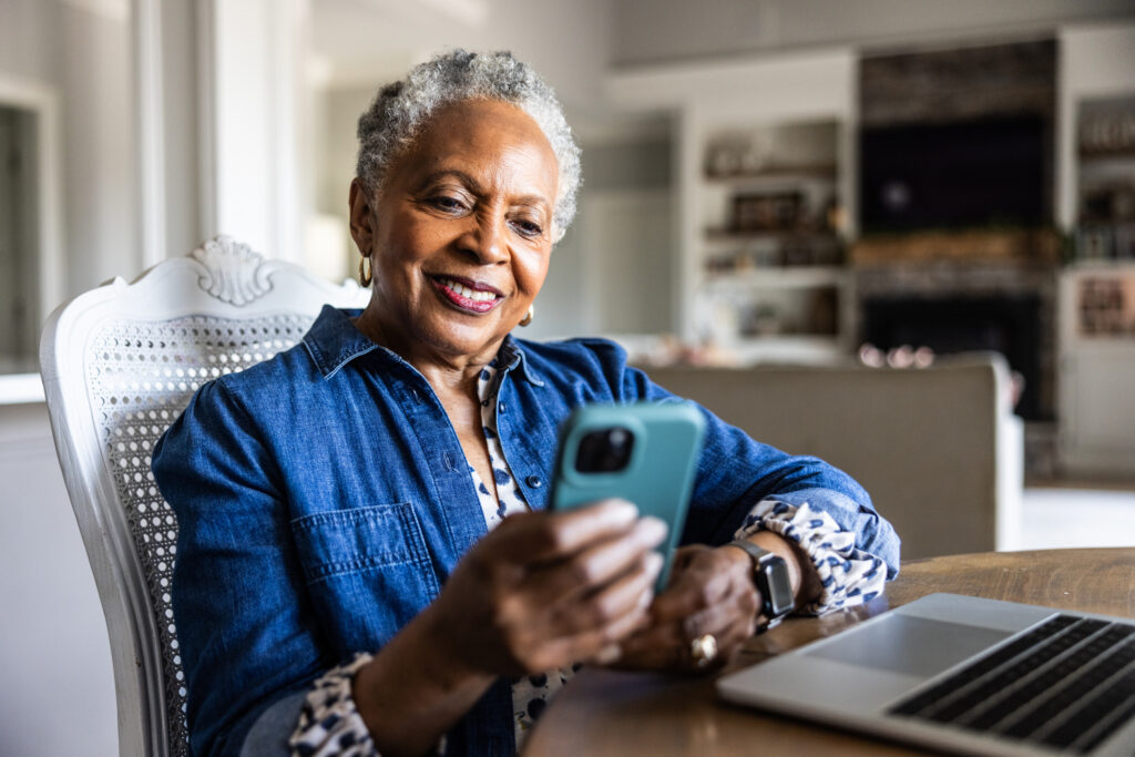 The Most Popular Apps for Retirement Planning in 2025 Older woman smiling and looking at cell phone while she sits at dining room table.
